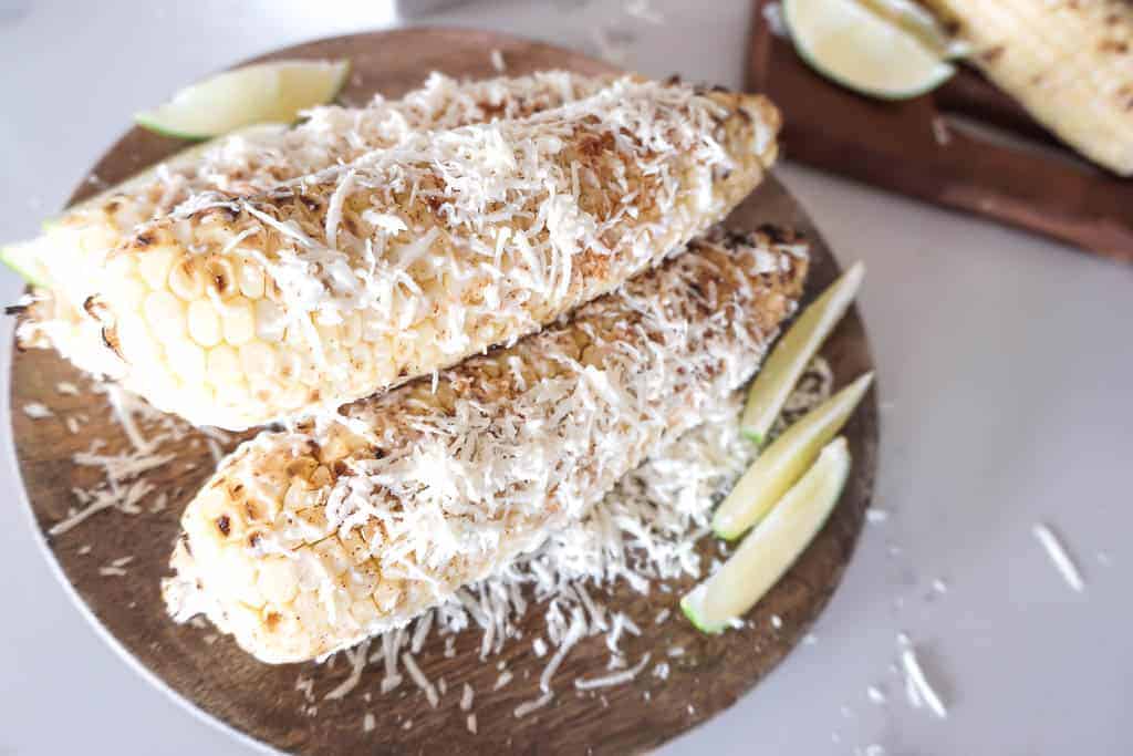 Mexican Street Corn pictured on a wooden plate with sliced limes. The Mexican Street corn is covered in chili powder and thinly grated cotija cheese