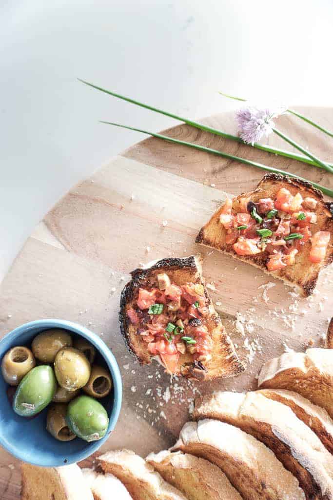 Signed Samantha's olive and bruschetta recipe - a birdseye view of olive bruschetta spread on a toasted baguette. There are two pieces placed on a cutting board with olives in a bowl sitting next to it.