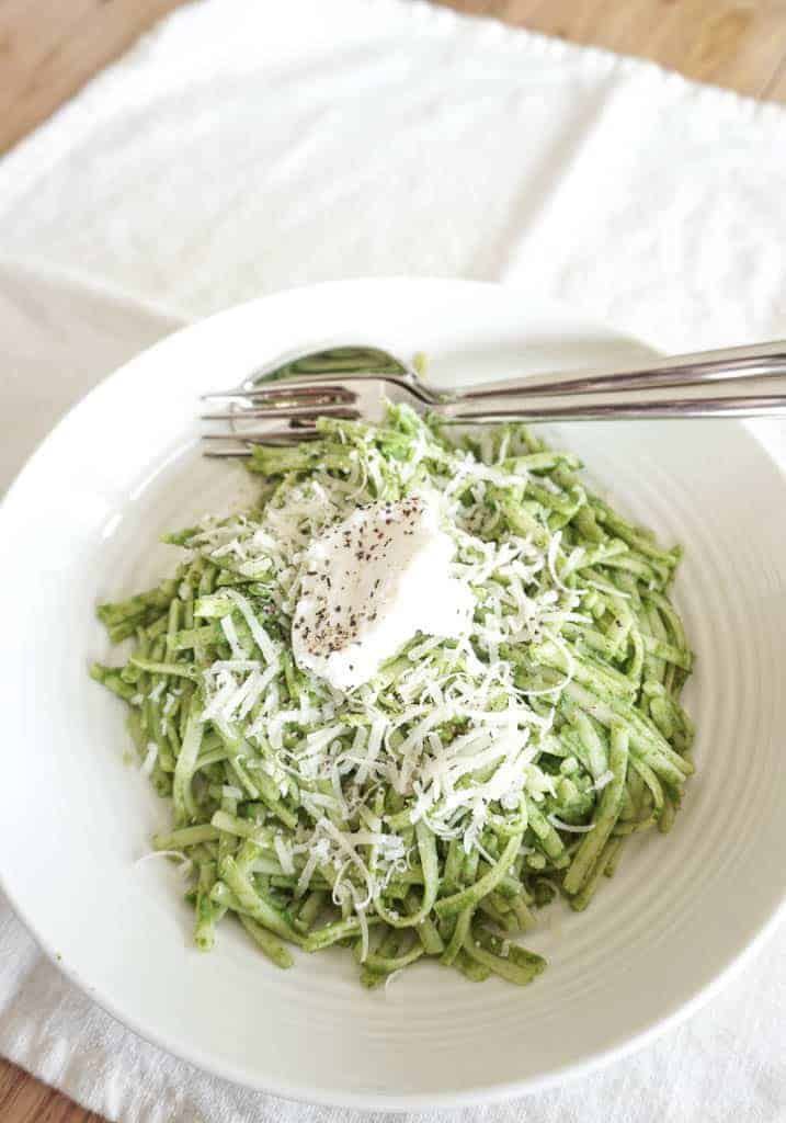 A finished look at the kale pasta recipe from Signed Samantha. In a low pasta bowl is green sauce on top of spaghetti. It is topped with parmesan cheese and ricotta cheese along with salt and pepper. There is a napkin under the bowl and a fork and spoon in the bowl.