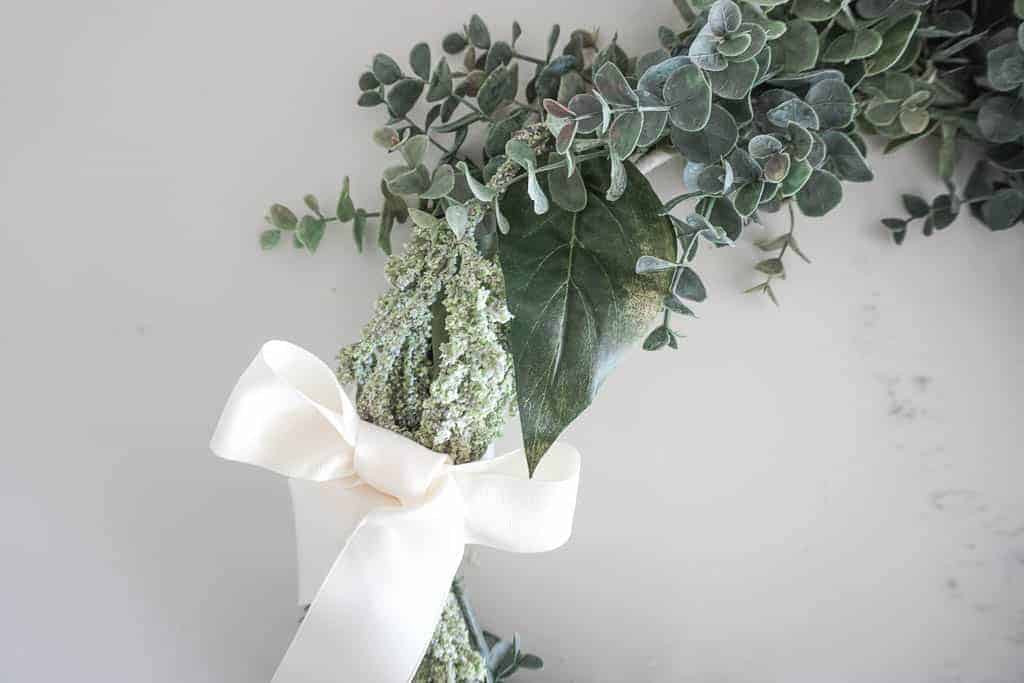 Signed Samantha's instructions on how to make a wreath step by step are included and limited items are needed. Pictured is 1/6th of the wreath with green eucalyptus, and another faux long green plant. The long green plant is tied in a bow with a cream coloured ribbon.