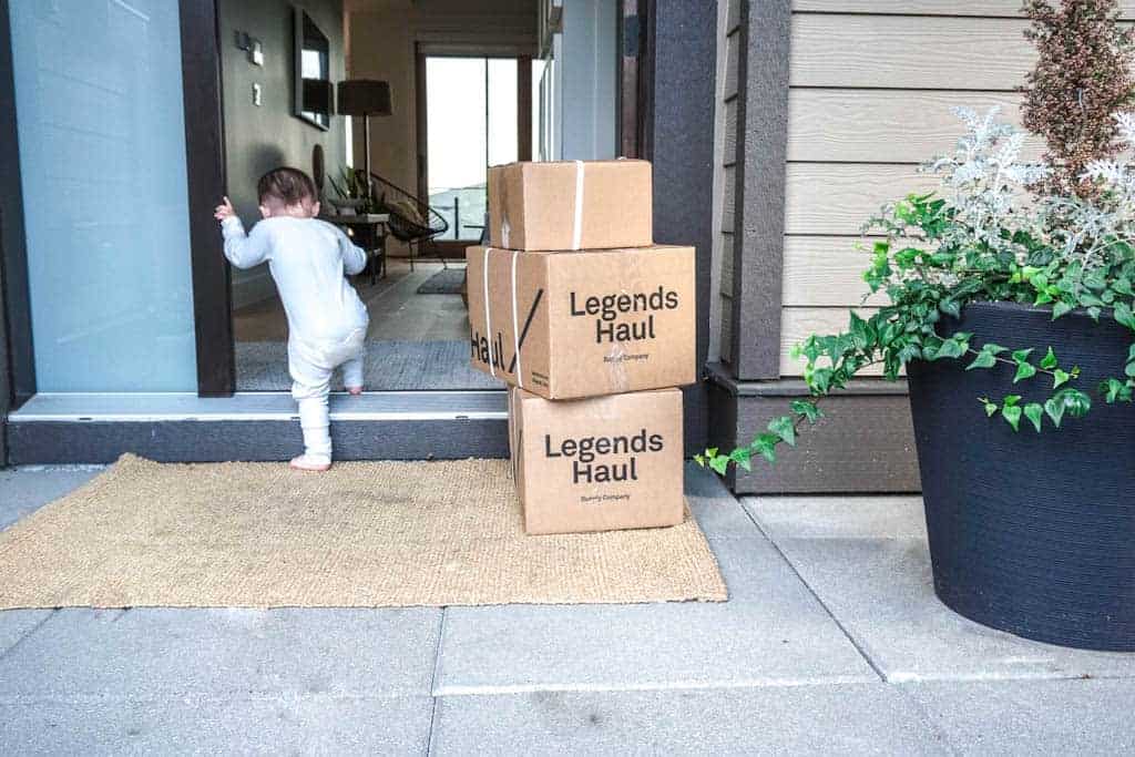 Signed Samantha's shop local Vancouver includes Legends Haul, a grocery delivery services. Three coardboard boxes with legends haul written on them are stacked outside the front door with a toddler running into the door.