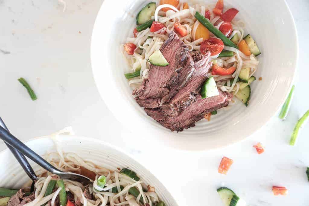 Signed Samantha's Rice Noodle Salad recipe picture which includes rice noodles, chopped cucumber, green beans, red peppers, and flank steak pictured in a white pasta bowl. Off to the side is the serving bowl of the noodles.