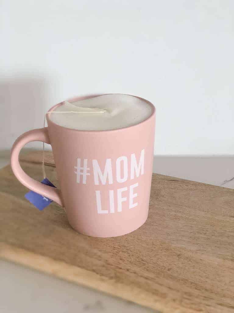 Tea time for Signed Samantha with a large, pink tea mug that says #momlife sitting on a wood cutting board. The mug contains a tea latte with beautiful foamy milk.