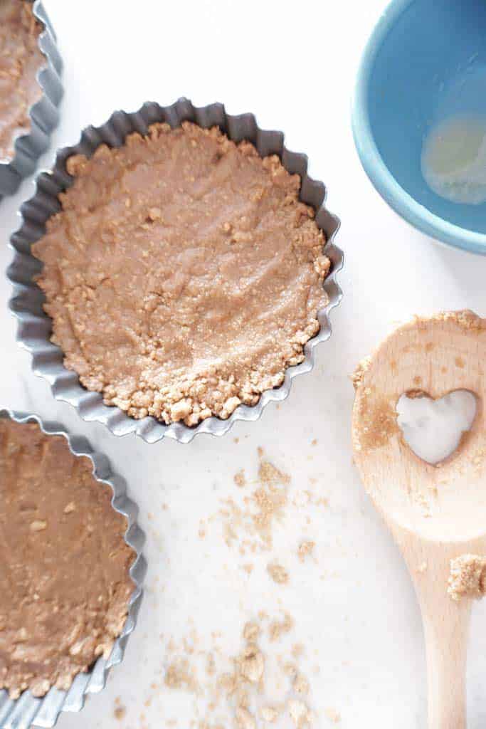 Signed Samantha's vegan gluten free s'more tart graham cracker crusts in mini 4 inch tart pans. There are two tart pans pictured filled with crust, and a wooden spoon and empty bowl in the image.