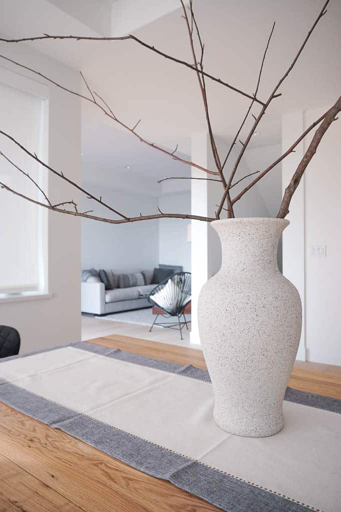 Signed Samantha's DIY stone vase is pictured on top of her dining room table which is a wood table with a runner on it. There are many large branches coming of the stone vase. In the background is Samantha's living room.