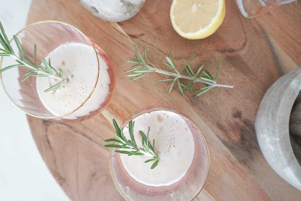 Signed Samantha's bourbon sours poured into two cocktail glasses each with a spring of rosemary in them. In the background there is rosemary, lemon, and a motor and pestle