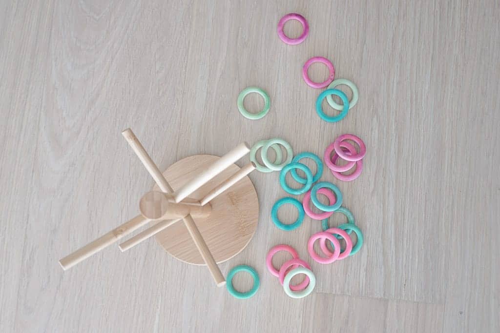 D.I.Y Ring Toss for Toddlers includes dollar store rings that are coloured different colours that get placed on a wooden mug holder. The wooden mug holder is on the ground and the rings are spread out around it.
