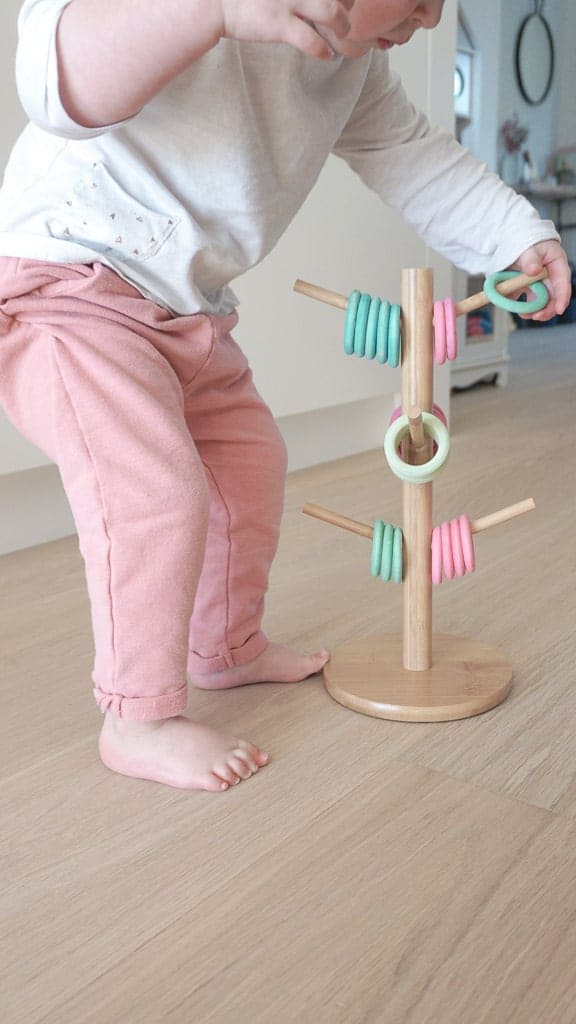 D.I.Y Ring Toss for Toddlers includes dollar store rings that are coloured different colours sitting on a wooden mug holder. Signed Samantha's daughter is standing putting a ring on one of the arms of the mug holder.