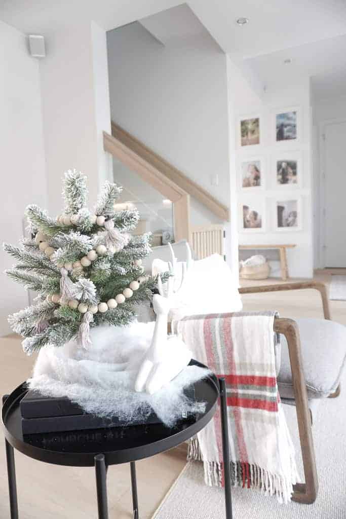 Signed Samantha run's through her 2020 Holiday Decor Wish List and vibe, which includes greens, and neutral colours. Pictured is her chair with a plaid (light green and light red) blanket draped off the side, with a faux fur pillow along the back. On the end table is a white reindeer and green mini christmas tree with wood beads as garland