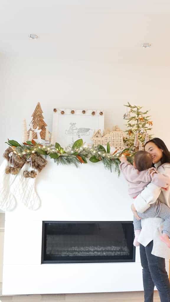 Signed Samantha sharing her holiday mantle decor ideas with you pictured is her mantle with garland and stockings in the front, trees, reindeer, and a mini tree behind. She is holding her daughter who is pointing at the decor.