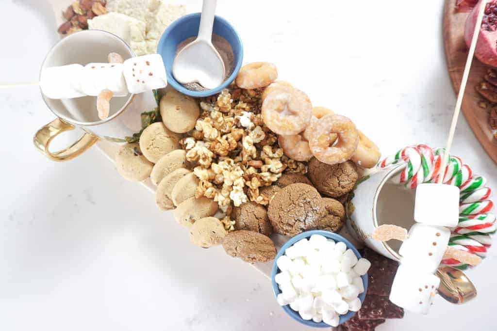 Christmas Grazing board ideas by Signed Samantha includes a sweets board her's has hot chocolate, mugs for hot chocolate, snowman marshmallows, cookies, and popcorn on it.