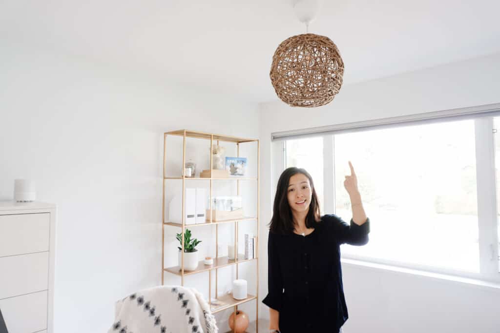 Signed Samantha standing below her D.I.Y pendant light in her home office which has a desk, laptop, and shelf in the background.