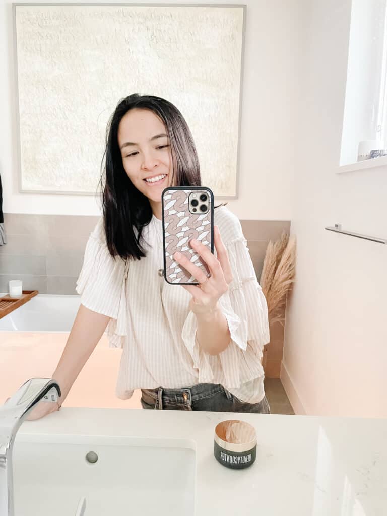 Signed Samantha standing in front of the mirror taking a selfie while teaching you how to use a cleansing balm, which is sitting on the counter (the cleansing balm).