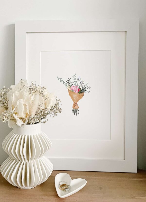 Signed Samantha's detailed bouqet print with light pinks and greens sitting in a frame next to a vase of dried flowers and a trinket holder with rings in it.