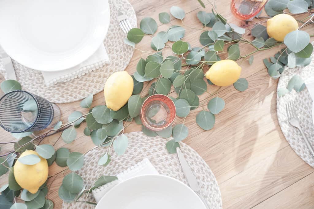Signed Samantha sharing some home decor updates for spring including how to update your tablescape for this season. Pictured is her wood table with eucaylptus and lemons as well as neutral napkins and placemats, white plates and coloured glassware.