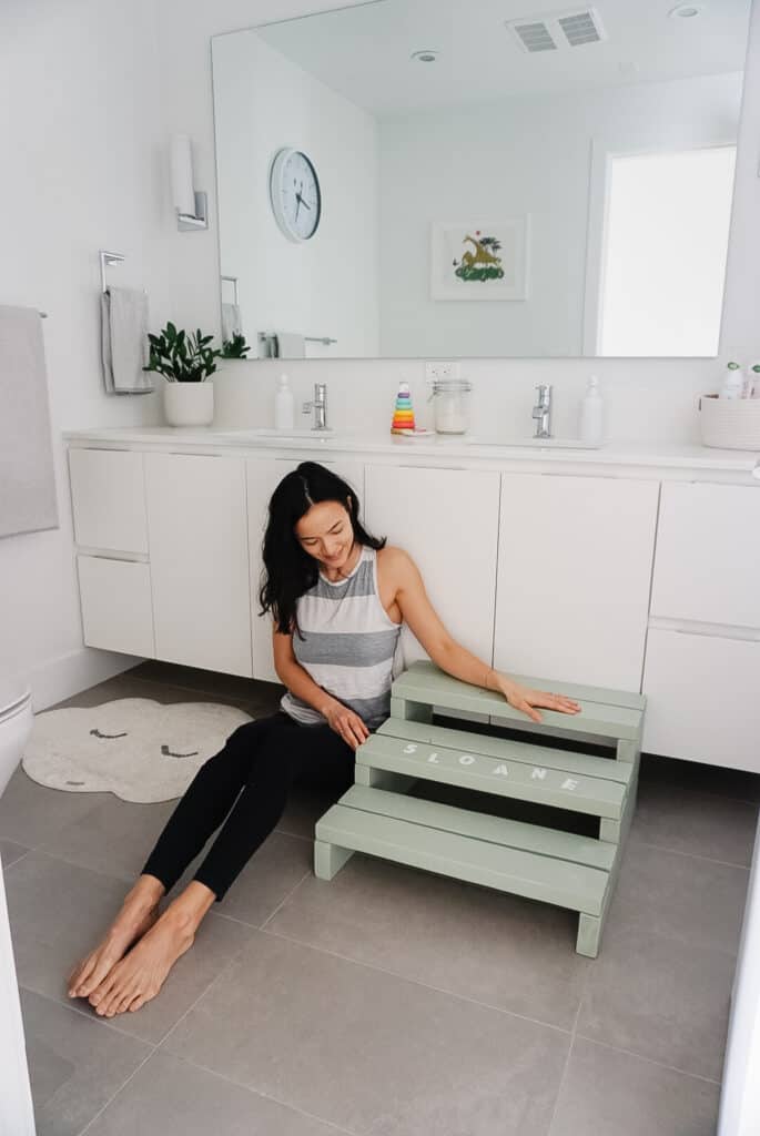 A sage green step stool for kids with Sloane's name on in it in white paint. Signed Samantha teaches you how to make your own step stool for kids. The stool is in a bathroom with two sinks and white cupboards and a big mirror. Samantha is sitting next to the stool admiring it.