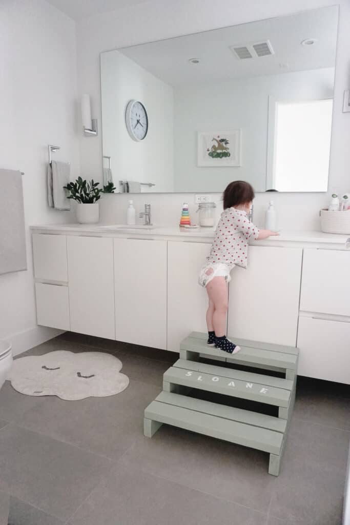 Signed Samantha teaching you How to Make Your Own Step Stool For Kids - Sloane is standing at the bathroom sink on hersage green step stool reaching the water.
