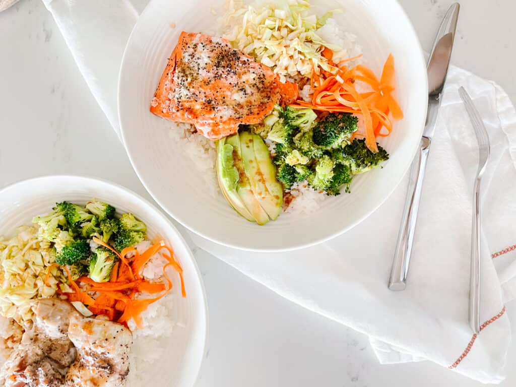 Salmon Rice bowls pictured from a birds eye view with salmon, rice, broccoli, avocado, and shredded carrots. There is a chicken option pictured next to the salmon bowl.