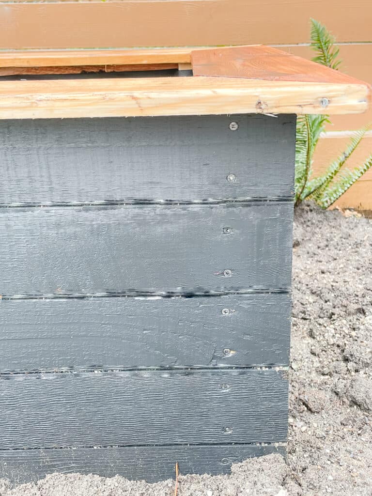 The corner of a garden box sitting outside in the garden bed. It is a black garden box with a non-painted cedar cap. this shows where the screw goes to get the bulk of the box together.