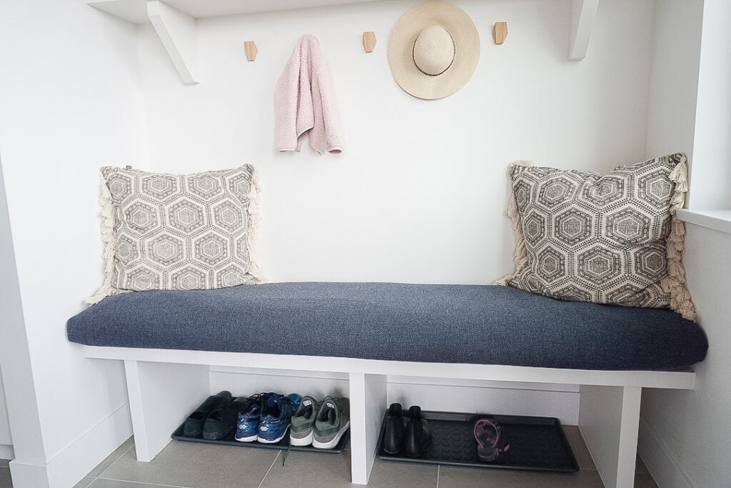 DIY Bench Cushion Cover in Samantha's mudroom - it is a blue thicker fabric material with throw pillows and a toddler jacket and hat on the hooks.