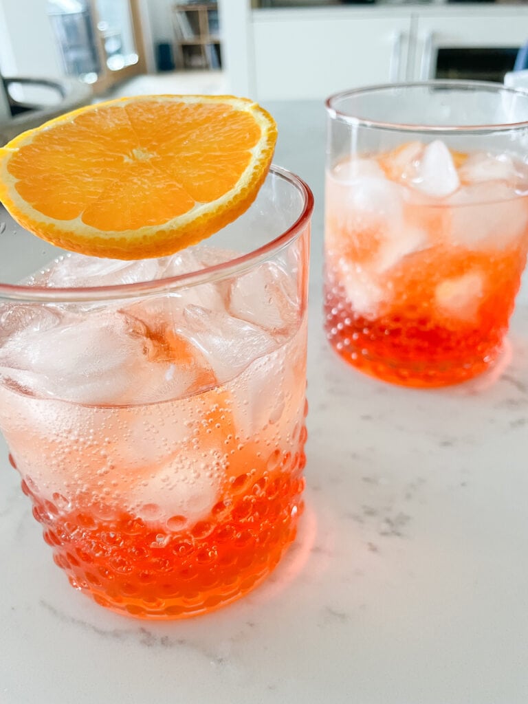 Aperol Spritz pictured on a white countertop. With orange garnish. The aperol spritz is in a pink glass.