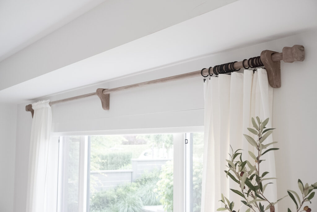 DIY Rustic Curtain Rod is pictured in signed samantha's living room. it is wooden with white curtains. The rod is stained very rustic-like.