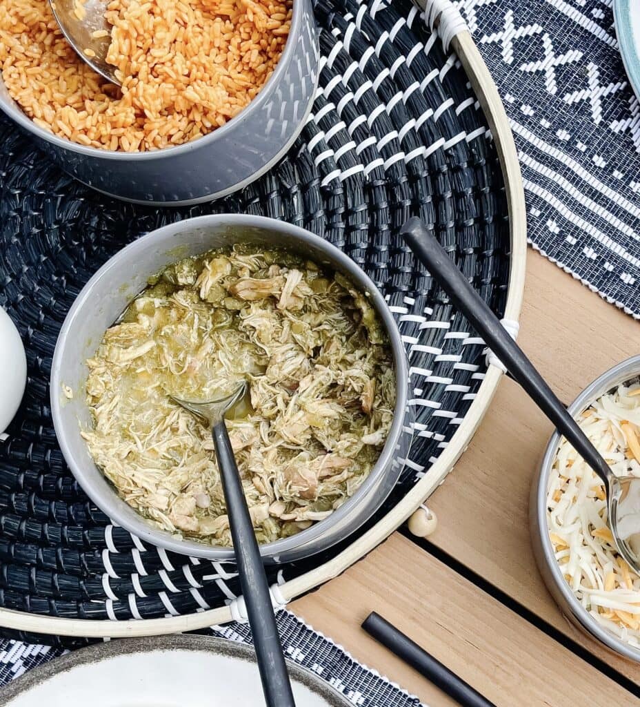 Slow cooker chicken verde pictured in a bowl on a table next to a bowl of shredded cheese and rice.