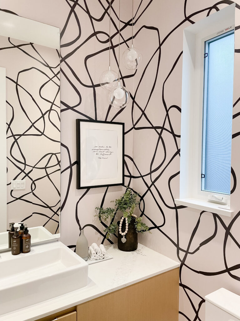 Peel and stick wallpaper in an abstract pattern on the wall - the background is a pinky taupe and the patterns are black. There is a black picture frame on the wall, a vase on the counter in the bathroom, soap, and towels.