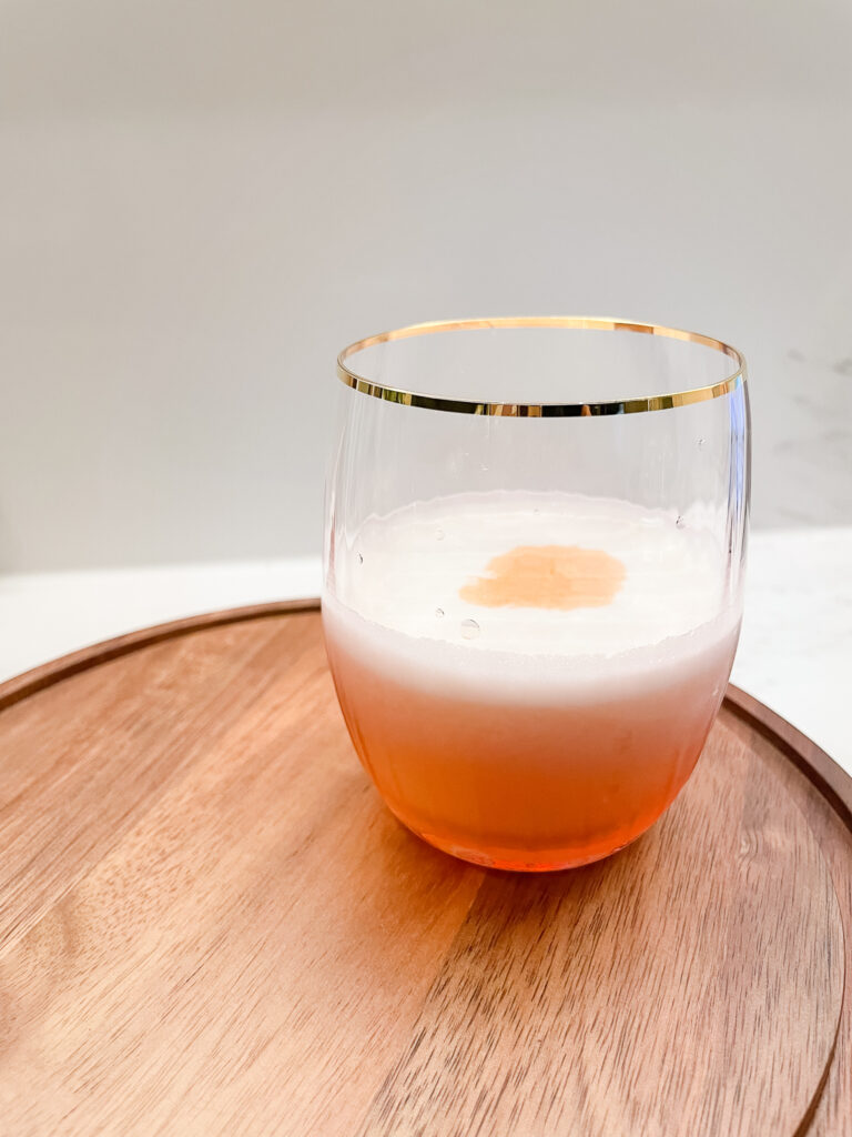 A smoked whiskey sour in a glass sitting on a wood board. There is a foamy egg white top with bitters topping it off.
