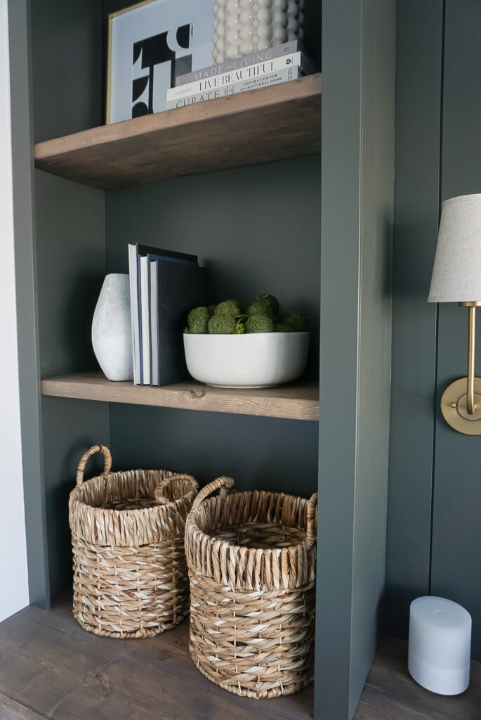 DIY Built-In Bookshelf in a green colour with shelves stained in a darker wood tone. The built-in bookshelf is sitting on top of a wood desktop. Decorated with baskets on the lowest shelf, bowls, books, plants, and pictures above.