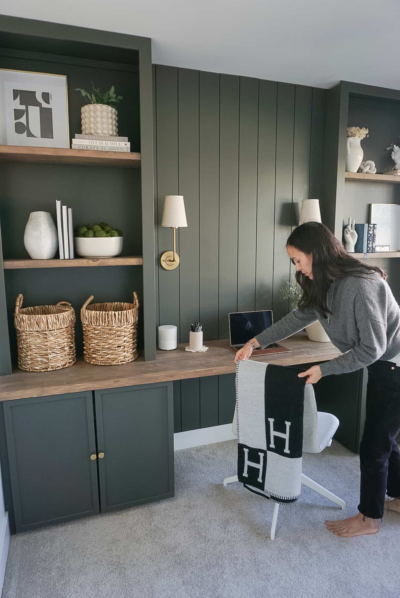 built in home office set up in a deep mossy green with shiplap and lighting at the back, bookshelves on either side and neutral decor throughout. Signed Samantha is standing fixing the blanket on the office chair.
