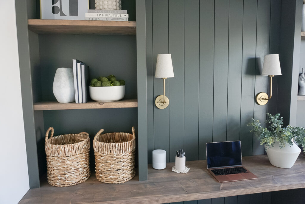 built in home office set up in a deep mossy green with shiplap and lighting at the back, bookshelves on either side and neutral decor throughout.