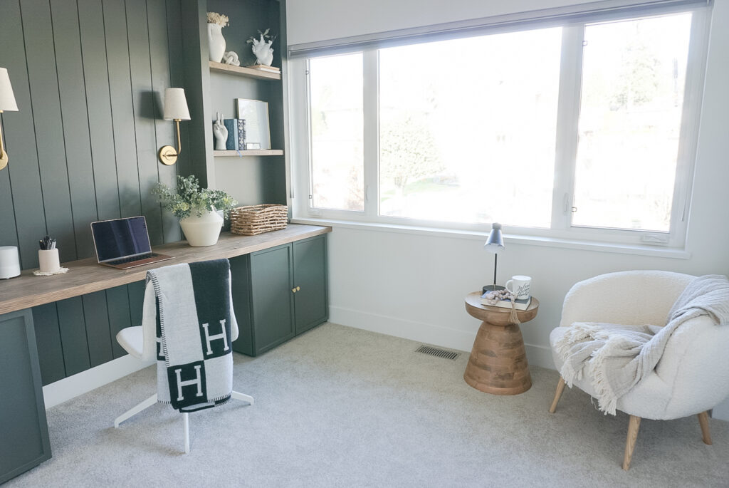 built in home office set up in a deep mossy green with shiplap and lighting at the back, bookshelves on either side and neutral decor throughout. A little nook in the corner with a chair and a end table are present.