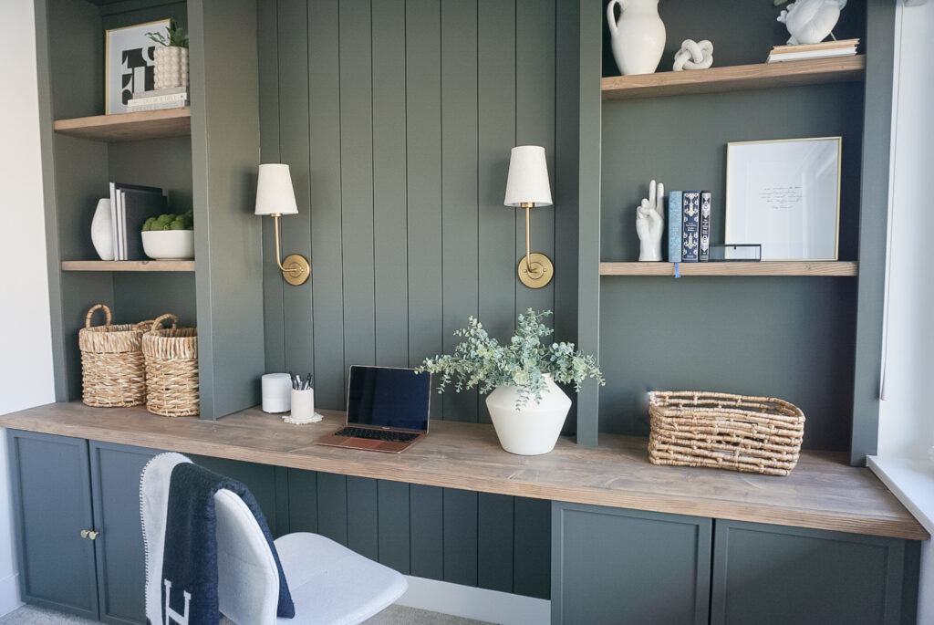 built in home office set up in a deep mossy green with shiplap and lighting at the back, bookshelves on either side and neutral decor throughout.