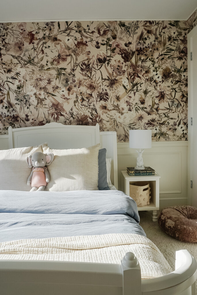 A big girl room with floral wallpaper, chair rail, box moulding, a white bed frame, blue duvet cover, end tables, and a pouf on the ground