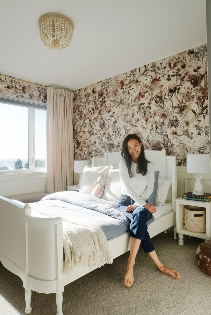 things to consider when costing a DIY project - A big girl room with floral wallpaper, chair rail, box moulding, a white bed frame, blue duvet cover, end tables, and a pouf on the ground plus signed samantha sitting on the bed.