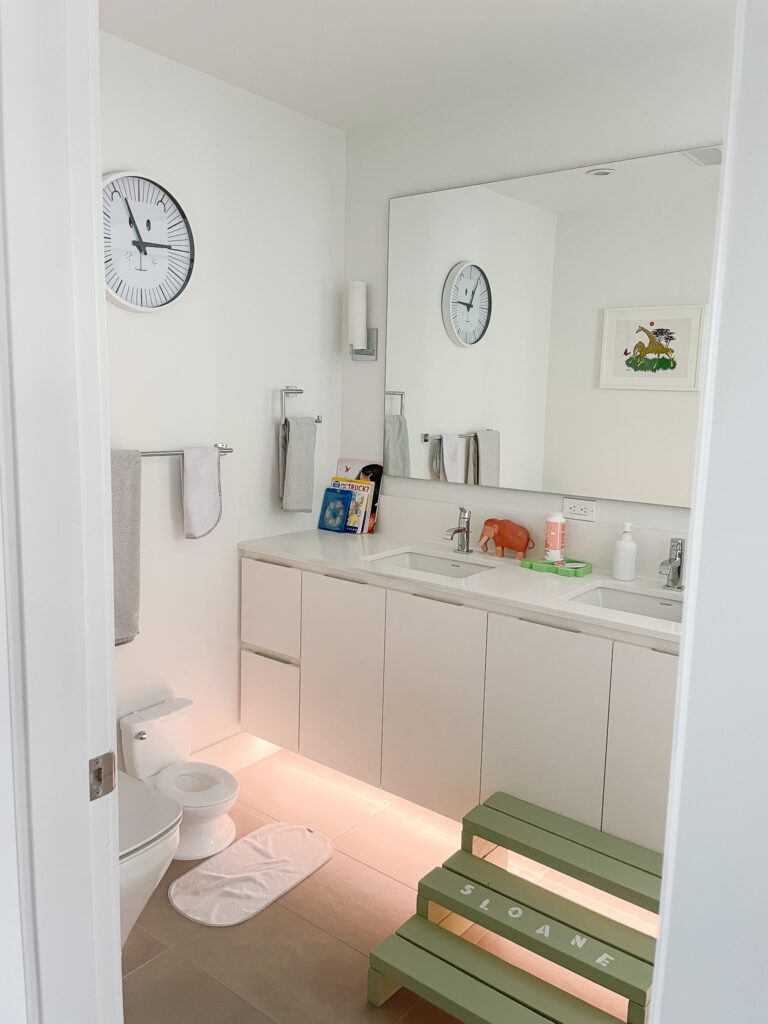 a builder grade bathroom with a full builder grade mirror, silver sconces, a stool and a bunch of children's books and toiletries on the counter