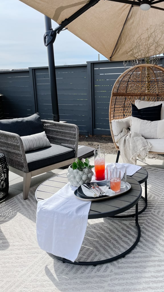 An outdoor patio oasis with two chairs, one egg chair and another club chair, a patio umbrella, outdoor coffee table, rug, and drink and snack set up.