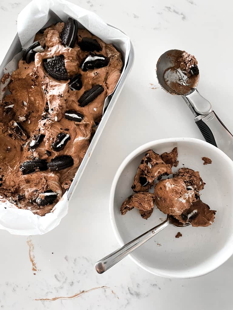 chocolate oreo ice cream pictured in a loaf pan and some in a bowl with a spoon and a ice cream scoop next to it